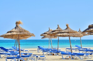 Een strand met rieten parasols en blauwe ligstoelen.