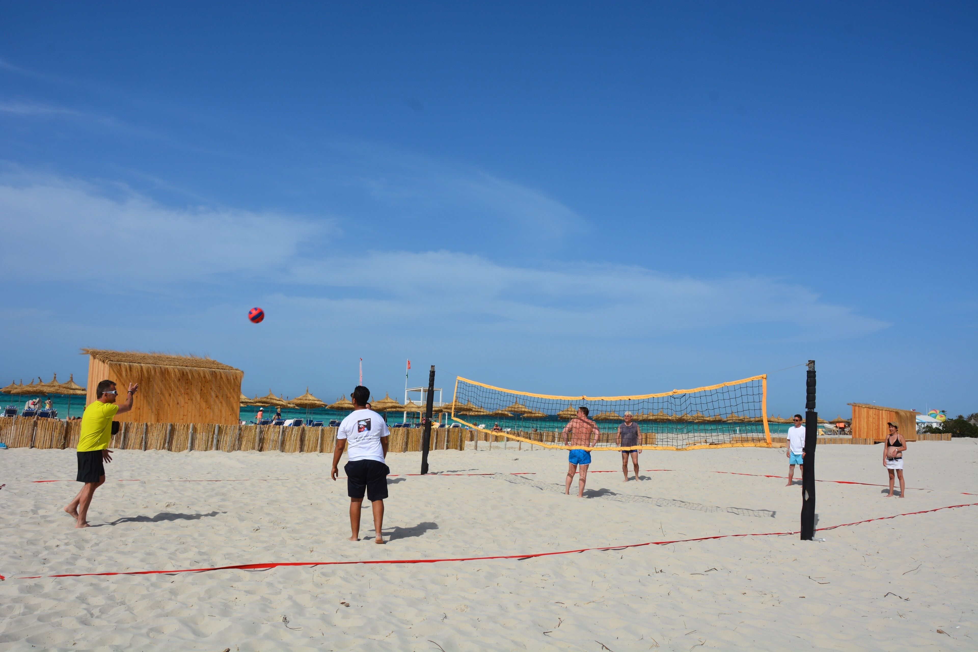 A beach volleyball game on a sandy court with a net, surrounded by beach huts and the ocean in the background.