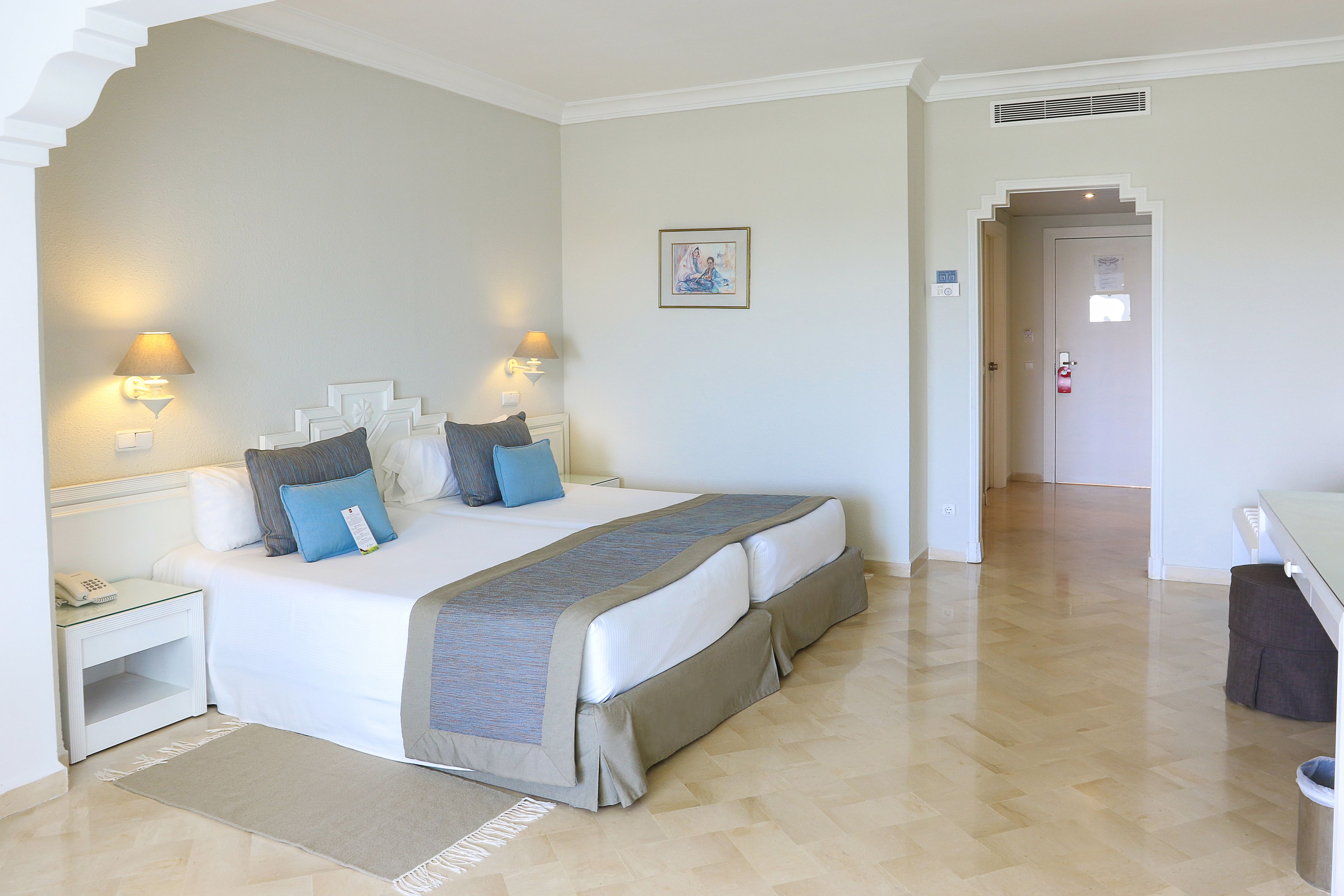 Una habitación de hotel con una cama grande, lámparas de noche, una mesita de noche, una alfombra y una puerta que da a otra habitación.