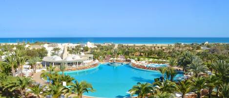 Un resort con una gran piscina central, palmeras y una playa al fondo.