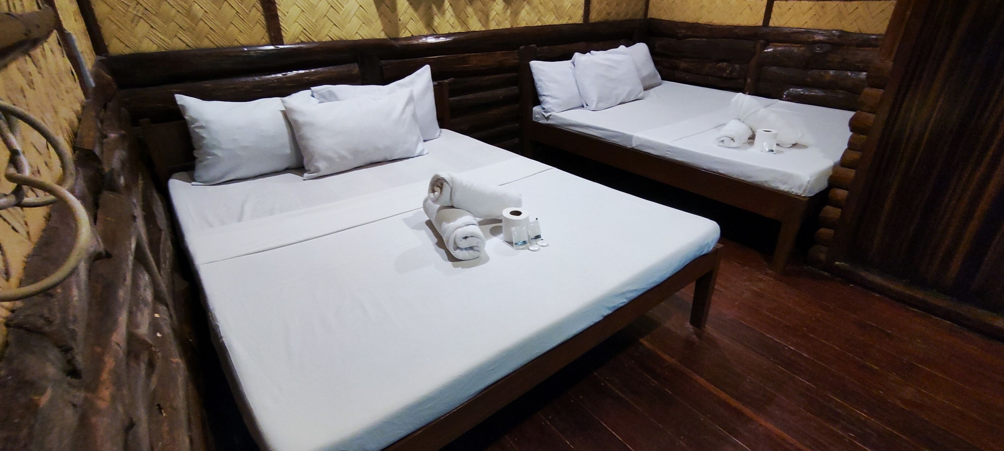 A room with two wooden beds, each with white linens and pillows. There are towels and toiletries on the beds.