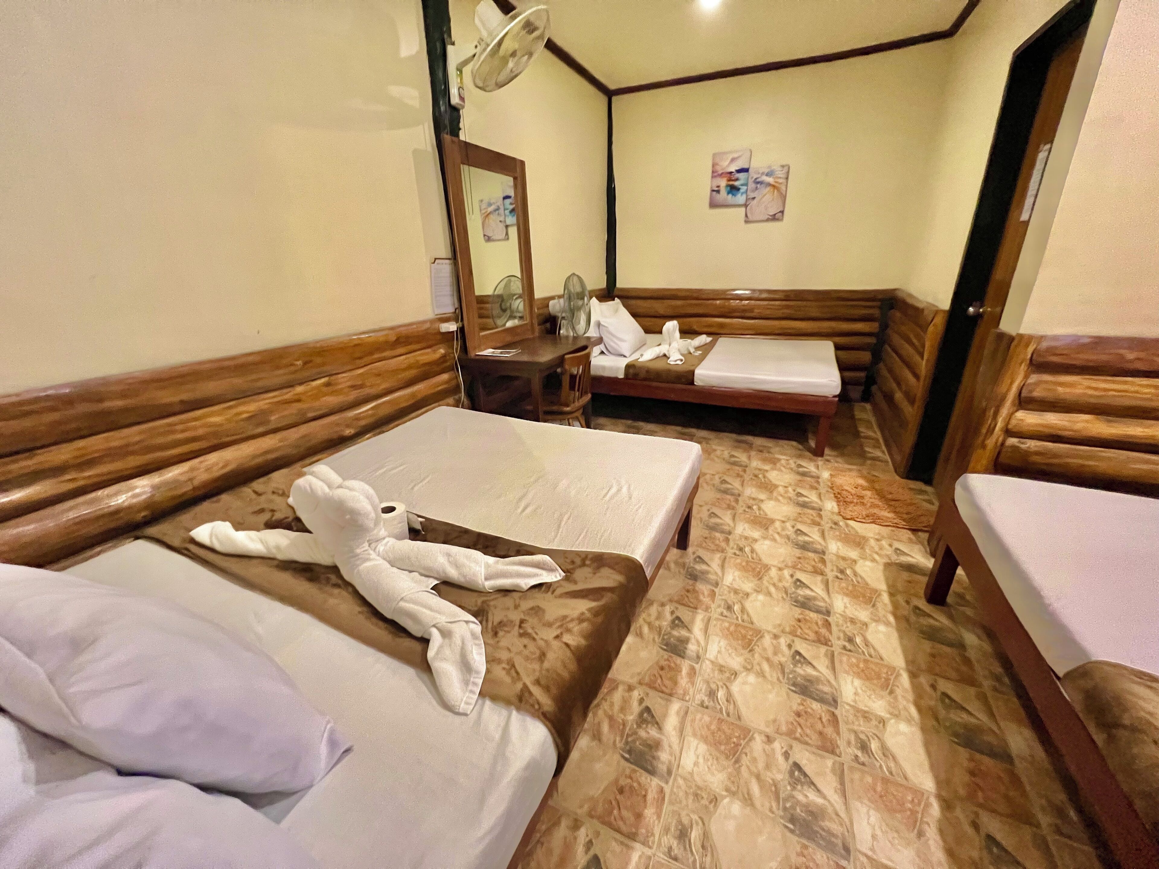 A hotel room with two beds, a wooden headboard, a mirror, a fan, and a tiled floor.