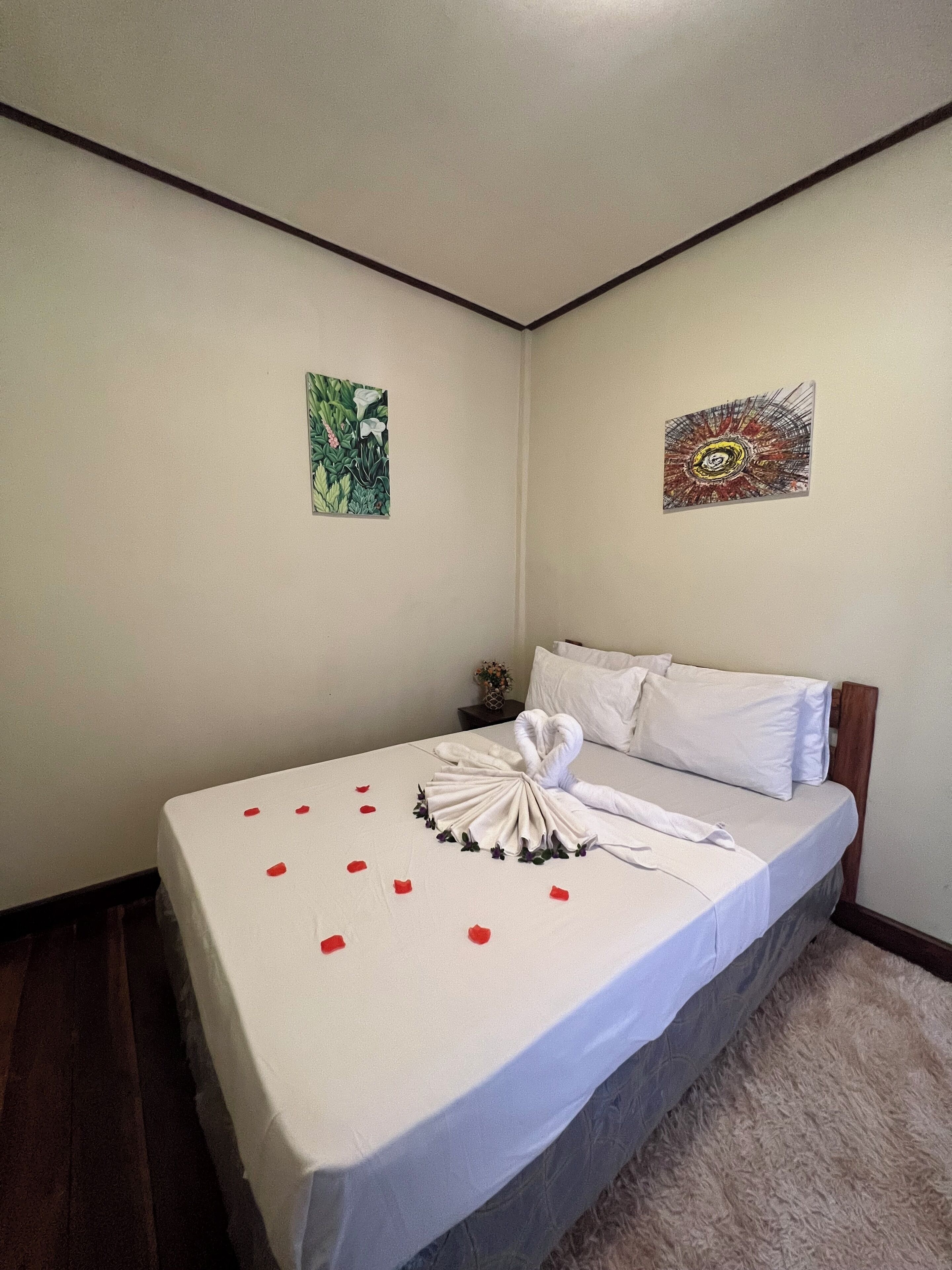 A neatly made bed with a towel arranged in a swan shape, red rose petals scattered on the bed, and two wall art pieces.