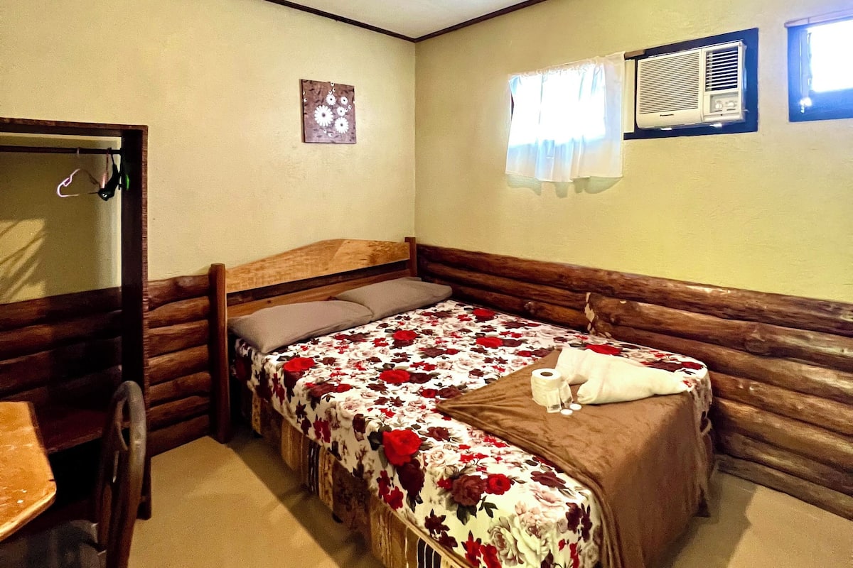 A room with a single bed, a wooden headboard, a small table, and a chair. There is a window with curtains, an air conditioning unit, and a ceiling light.