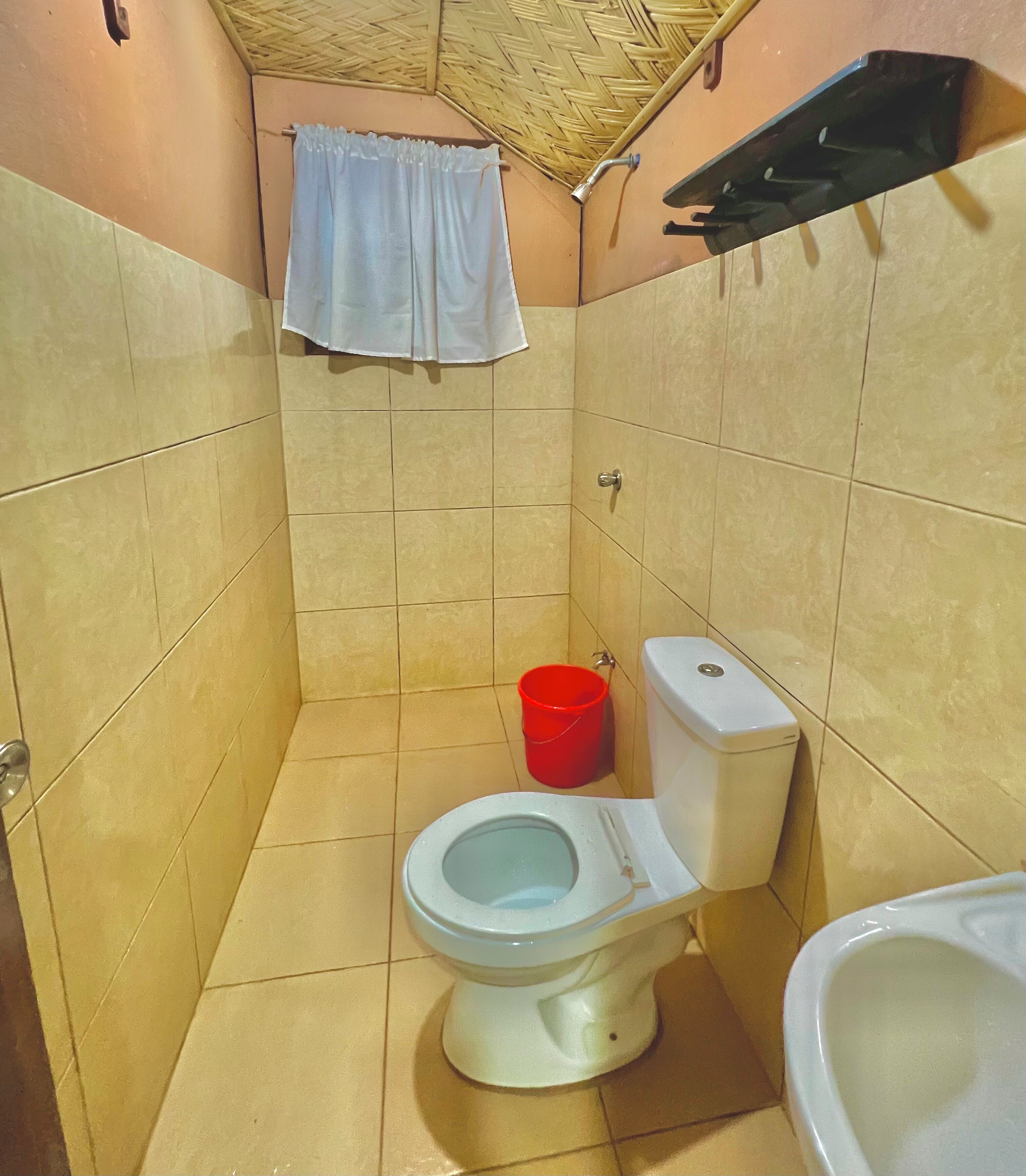 A small bathroom with a toilet, a red bucket, and a shower area.