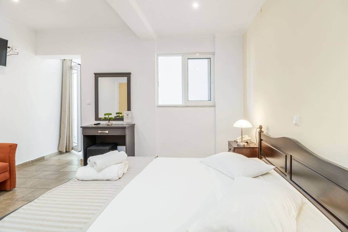 A modern hotel room with a large bed, a desk, a chair, a mirror, and a window.