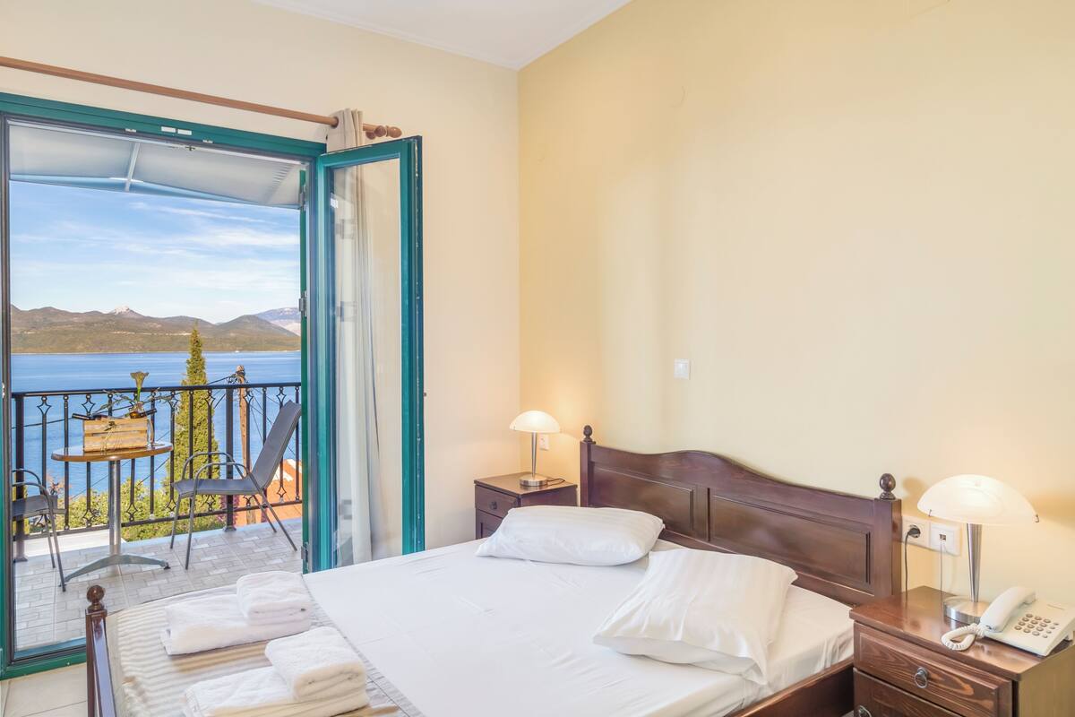 A hotel room with a bed, bedside tables, a balcony with a view of the sea and mountains.
