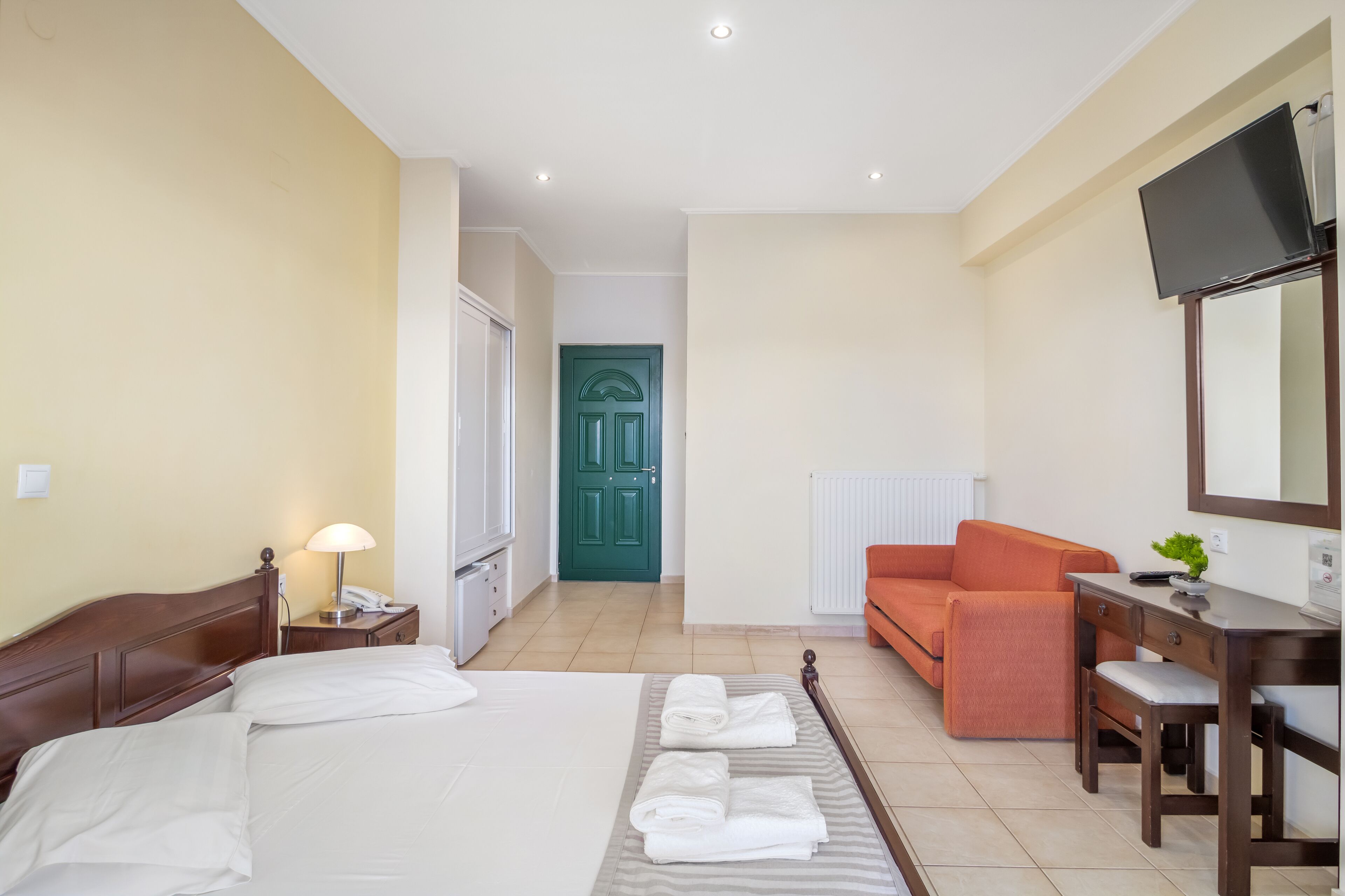 A hotel room with a bed, a desk with a chair, a television, and a green door.