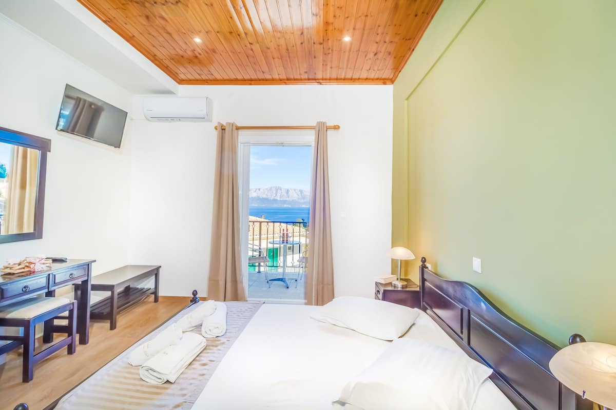 A hotel room with a wooden ceiling, a bed with white linens, a desk with a chair, a TV mounted on the wall, and a view of the sea through a window.