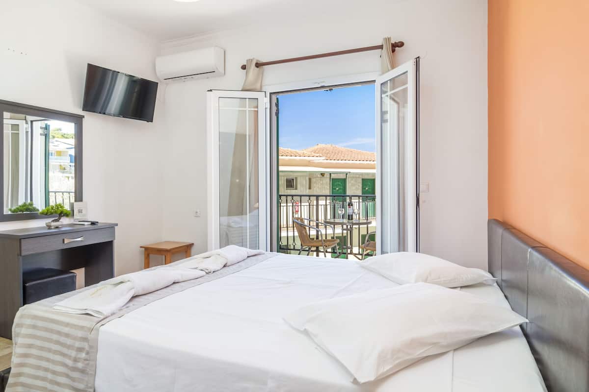 A hotel room with a bed, a desk with a chair, a TV mounted on the wall, and a balcony with a table and chairs.