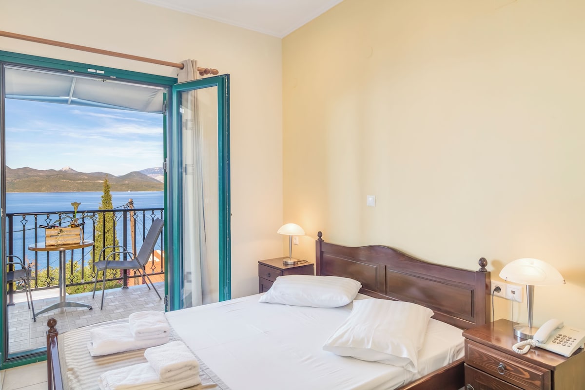 Habitación de hotel con cama, mesitas de noche y balcón con vistas al mar y a las montañas.
