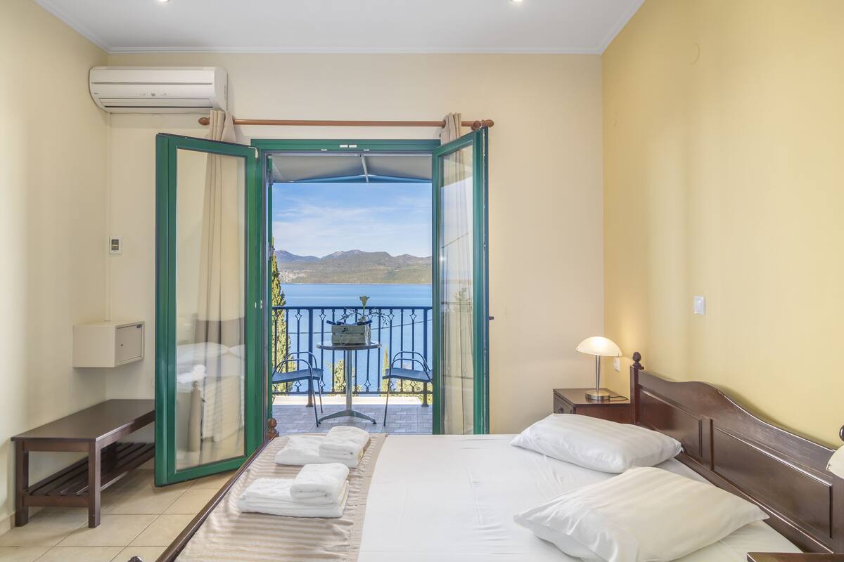 A hotel room with a bed, a bedside table, a lamp, an air conditioning unit, and a balcony with a view of the mountains.