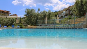 A swimming pool with clear blue water, surrounded by lounge chairs and umbrellas, with buildings and trees in the background. - Vergina Star (Lefkada)