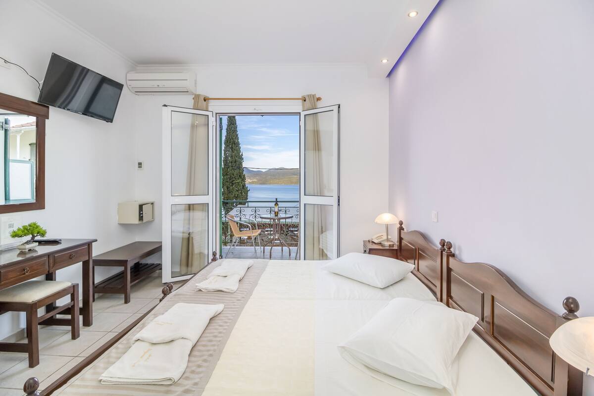 A hotel room with a bed, bedside tables, a desk, a TV, and a balcony with a view of the sea.