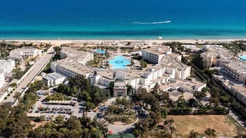 An aerial view of a coastal hotel with a central swimming pool, multiple buildings, and a beach in the background.