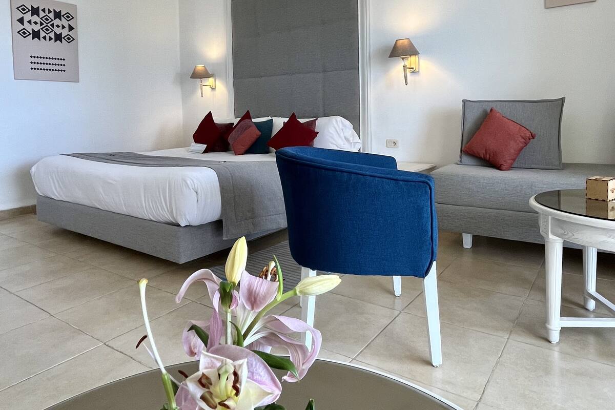 Ein modernes Hotelzimmer mit einem Bett, einer Couch, einem Sessel und einem kleinen Tisch. An den Wänden sind Lampen angebracht und ein dekoratives Element ist zu sehen.