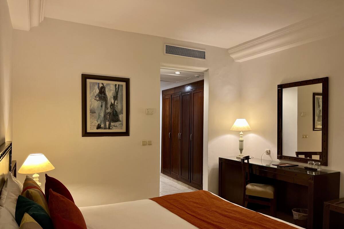 Ein Hotelzimmer mit einem Bett, Nachttischlampen, einem Schreibtisch mit Spiegel und einer Tür zu einem Flur.