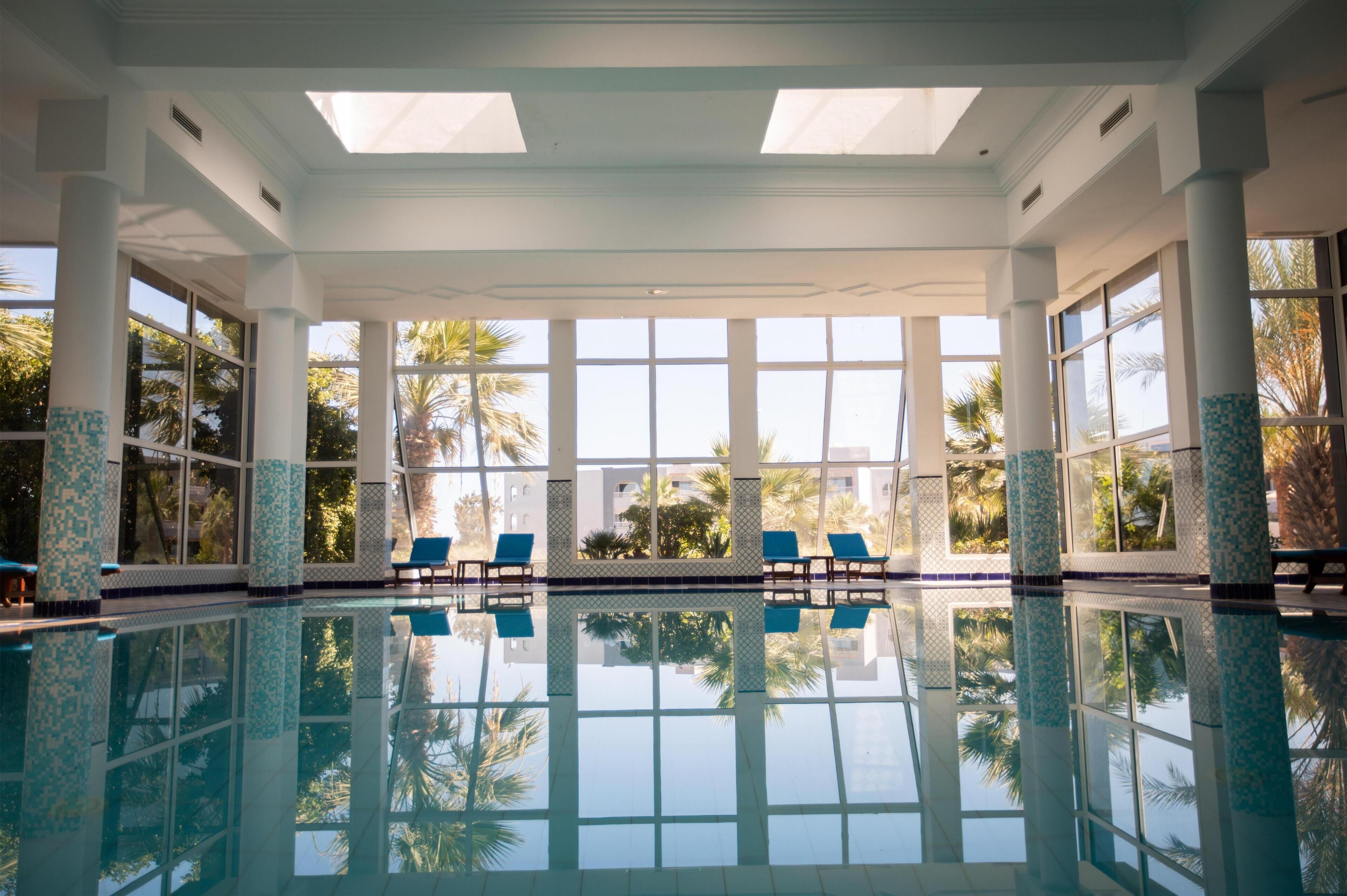 An indoor swimming pool with clear reflections, large windows, and blue chairs.