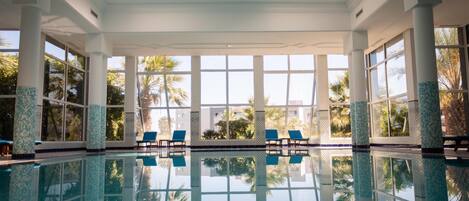 An indoor swimming pool with clear reflections, large windows, and blue chairs.