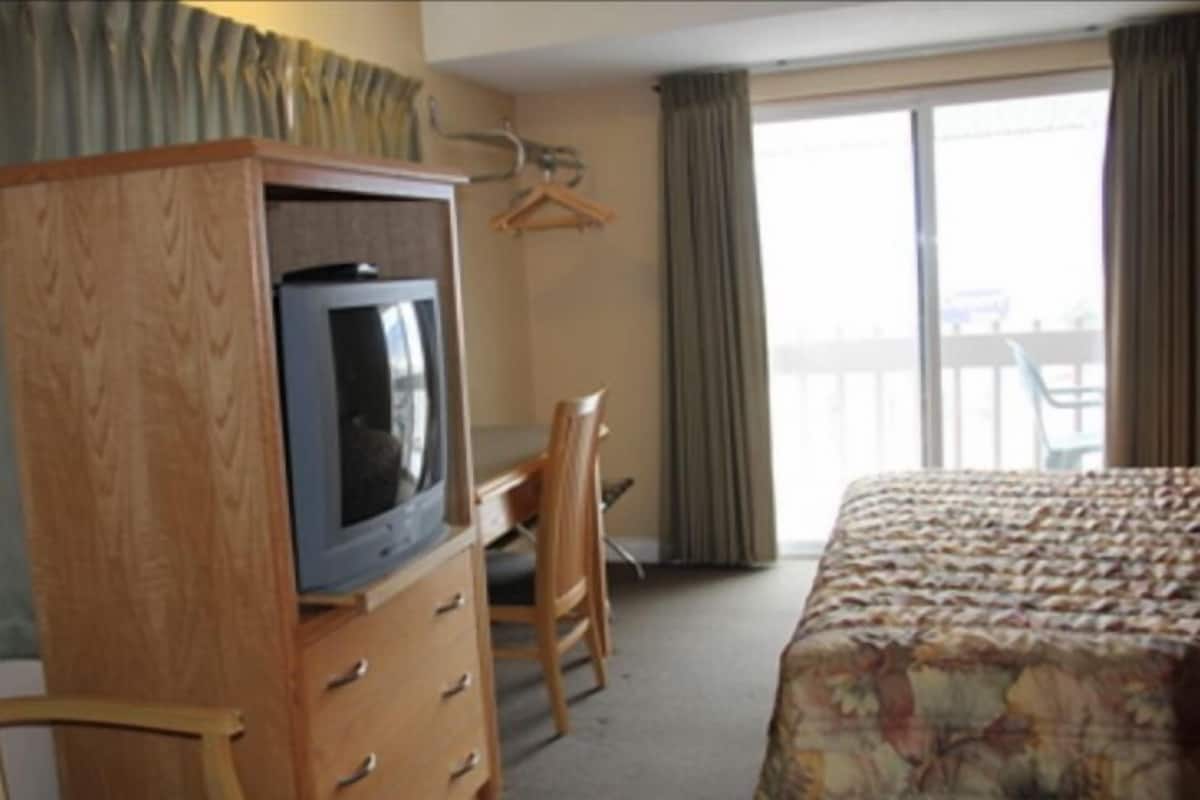 Une chambre d’hôtel avec une commode en bois, un téléviseur, un bureau, une chaise et un lit recouvert d’une couvre-lit à motifs floraux.
