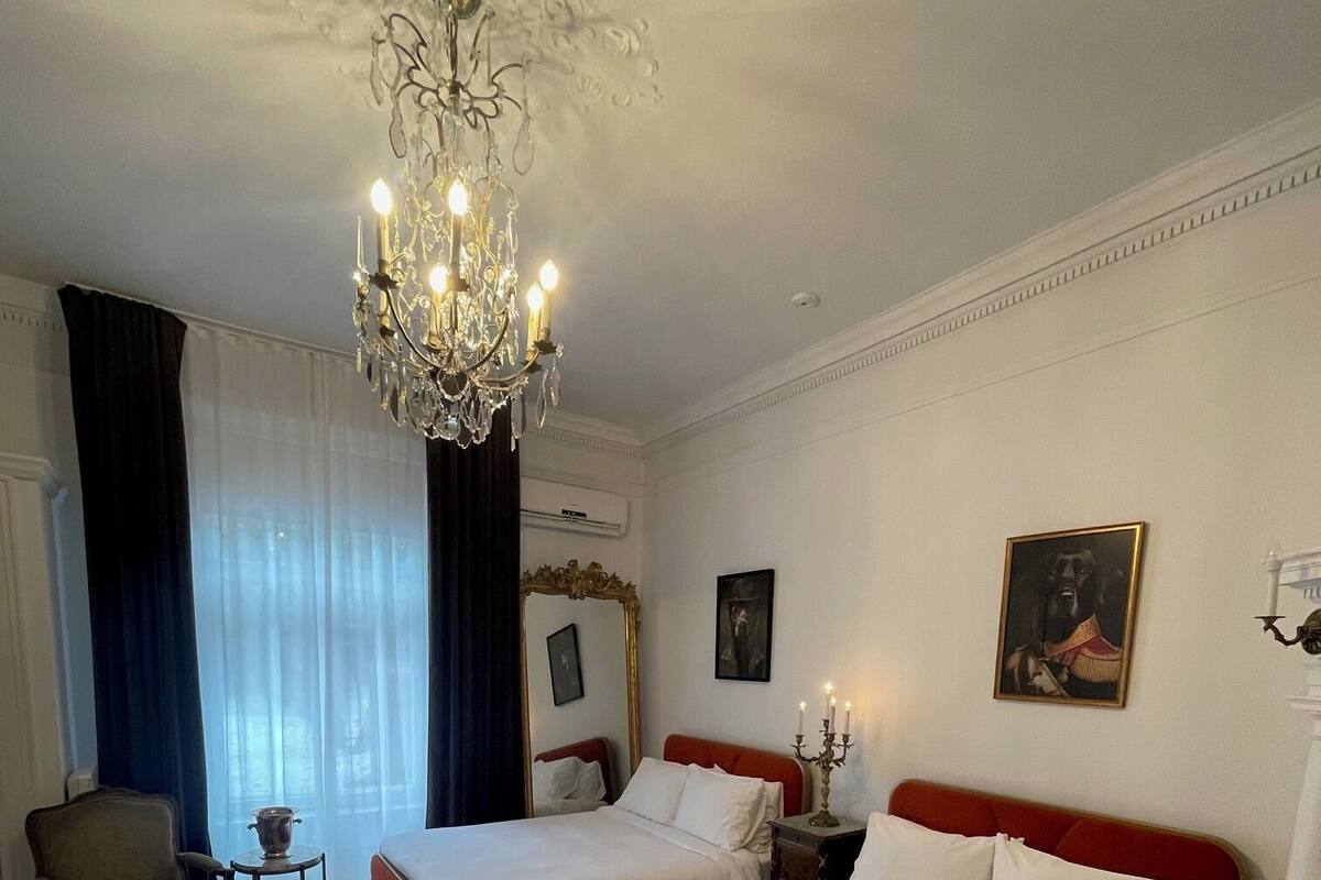 Quarto de hotel com duas camas, um candelabro, uma cadeira, uma mesinha e um quadro na parede.