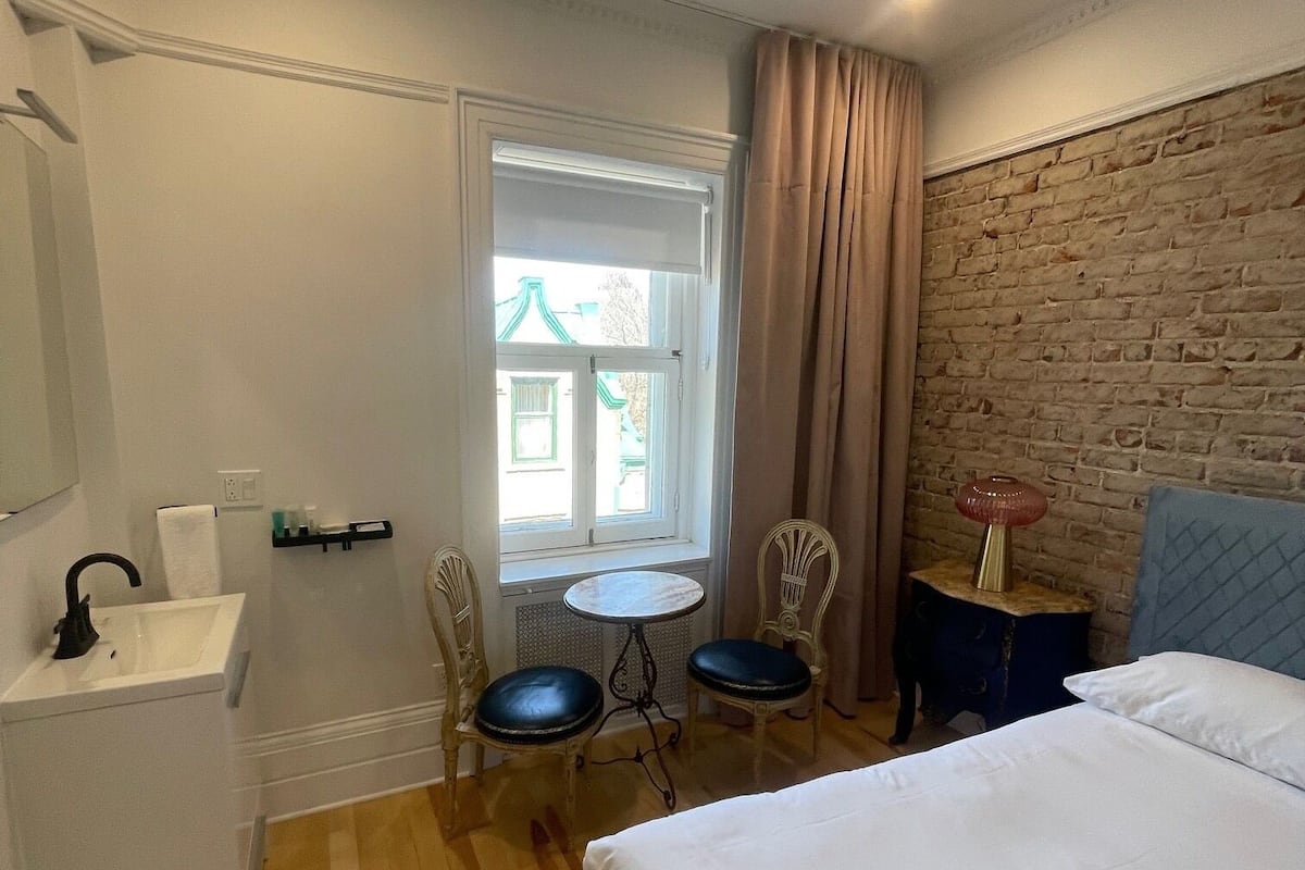 Quarto de hotel com uma cama, uma mesinha com cadeiras, uma pia e uma janela com cortinas.