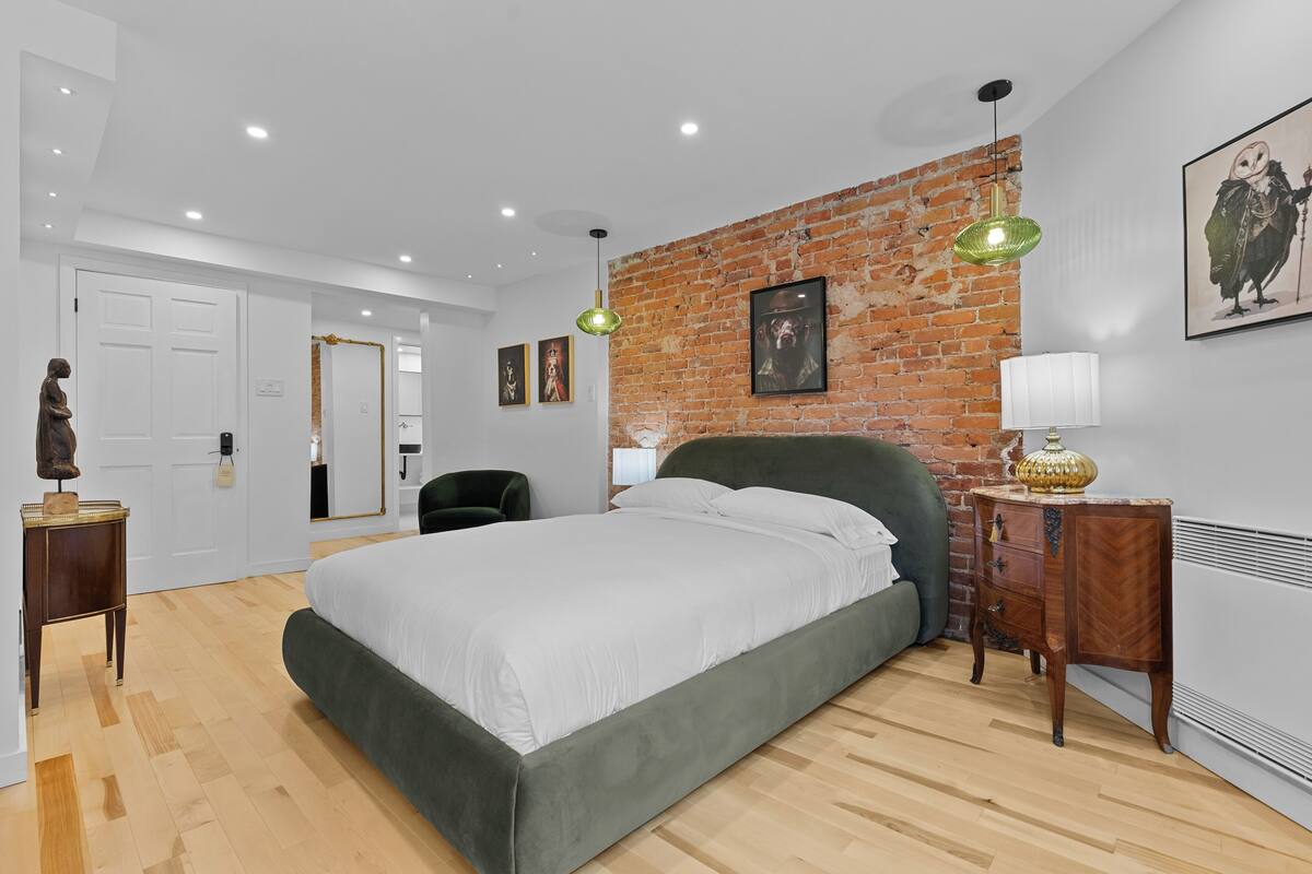 Quarto com cama grande, parede de tijolos, cabeceira verde, mesa de cabeceira de madeira e luminária pendente verde.