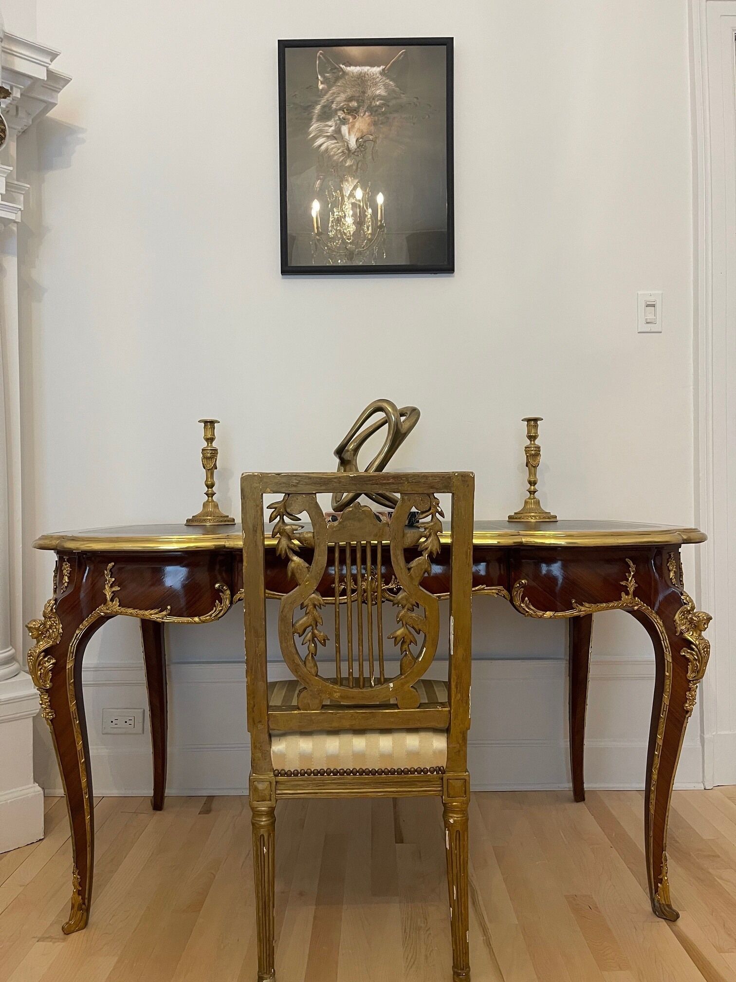 A room with a wooden floor, a dark wooden desk with gold accents, a gold-framed chair, and a framed picture on the wall.