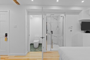 Una habitación de hotel moderna con piso de madera, cama, televisor, cabina de ducha de vidrio y un inodoro.