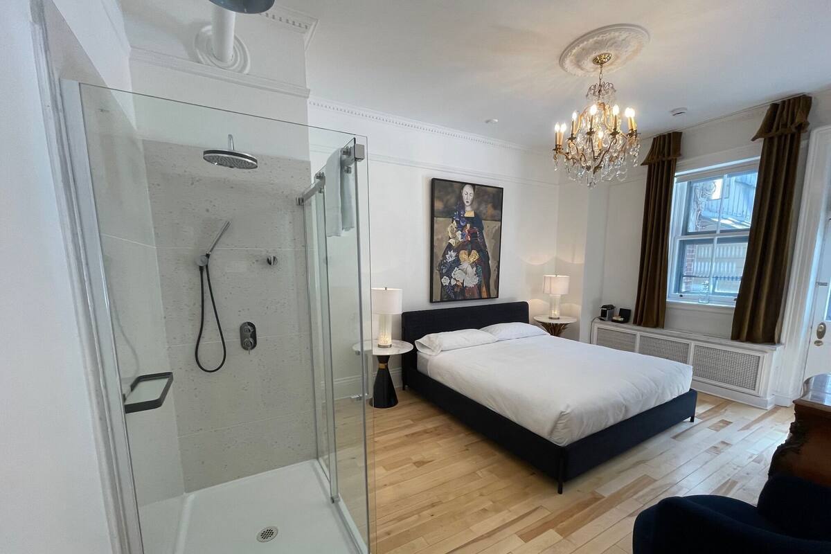 Quarto de hotel moderno com chuveiro em cabine de vidro, uma cama grande, uma mesa de cabeceira com abajur, um quadro na parede e um lustre.