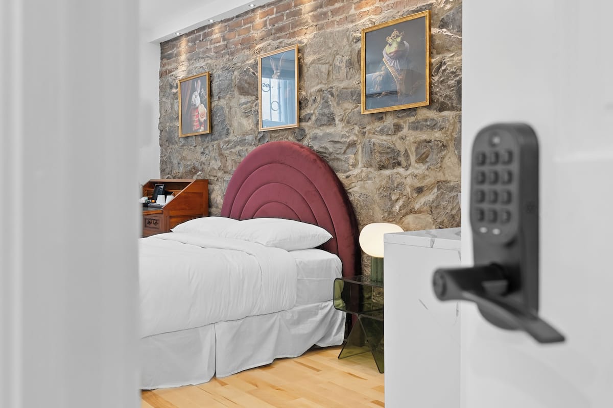 Quarto com parede de pedra, cabeceira vermelha, cama com roupa de cama branca, mesa de cabeceira e luminária.