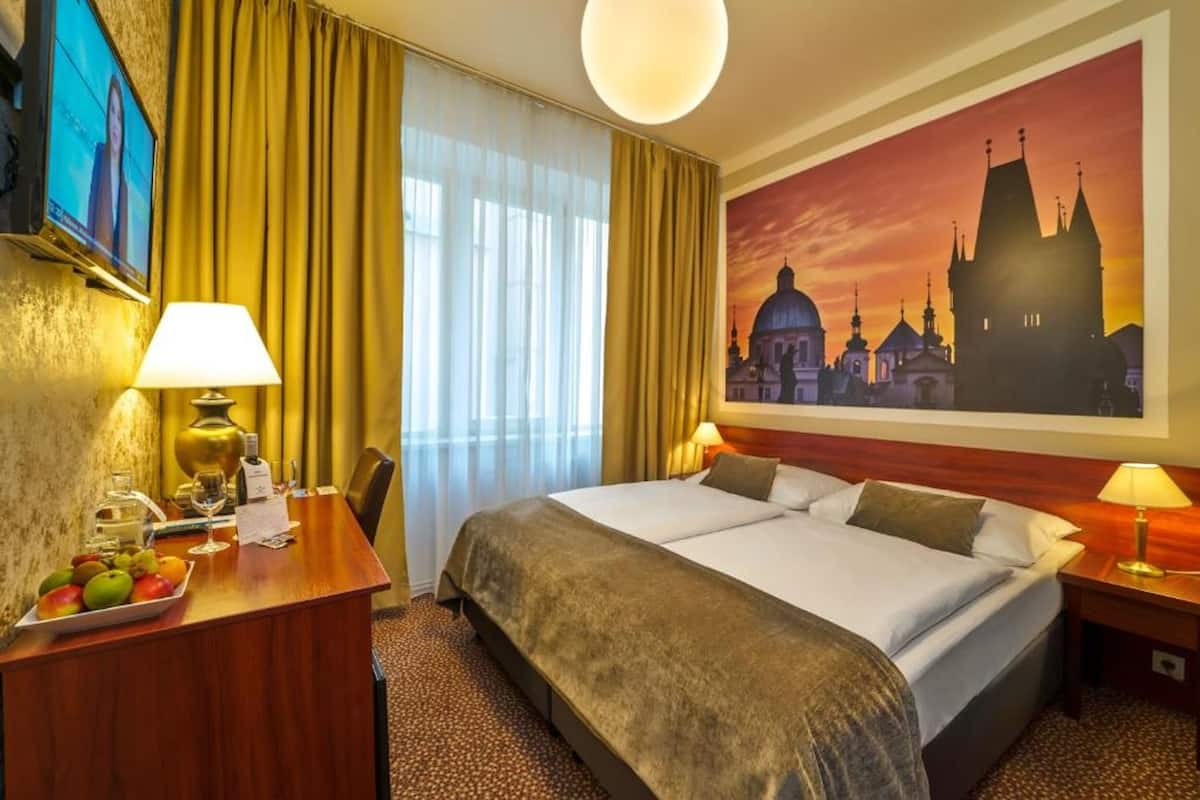 Een hotelkamer met een bed, twee bedlampen, een bureau met lamp, een schaal fruit en een schilderij van een stadsgezicht.