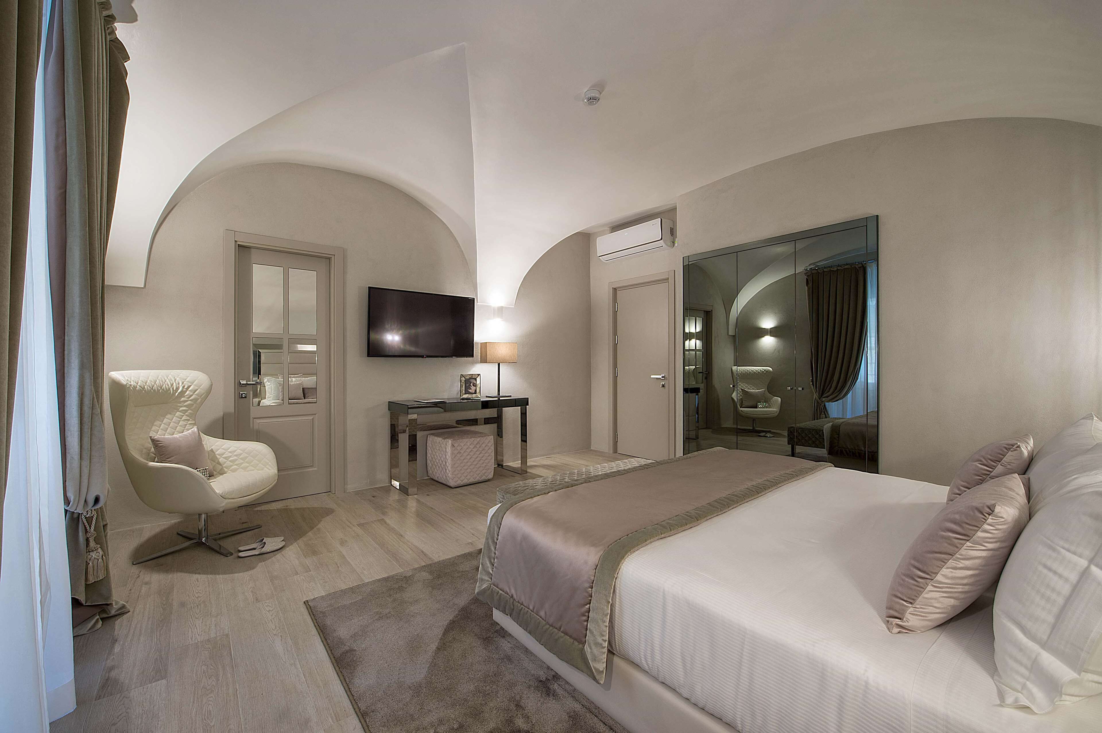 Quarto de hotel moderno com uma cama grande, uma poltrona, uma escrivaninha e uma televisão.
