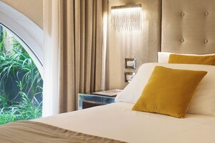 A hotel room with a bed, a nightstand, a lamp, and a view of greenery through a window.