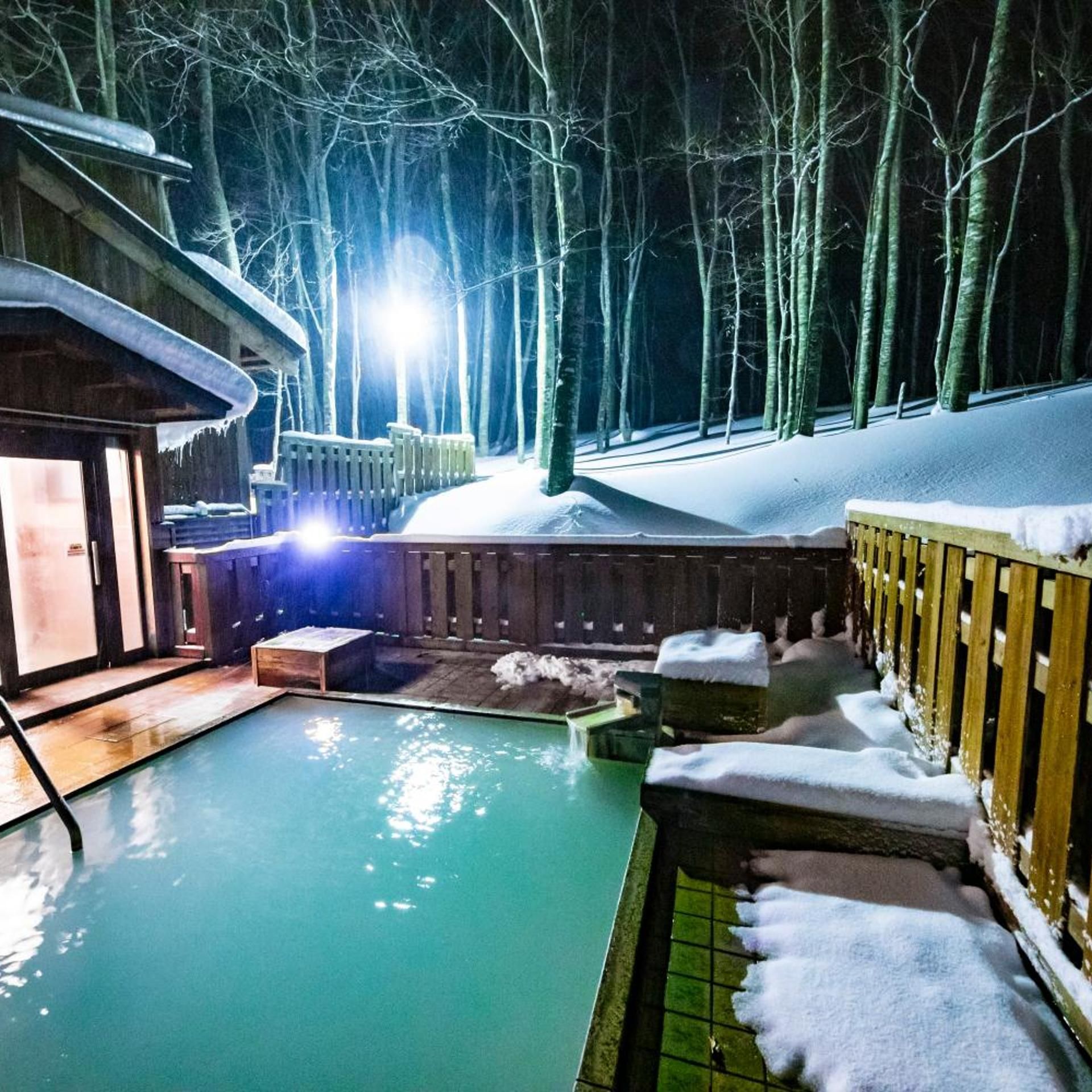 A heated outdoor pool surrounded by snow-covered wooden structures, with a forest in the background.