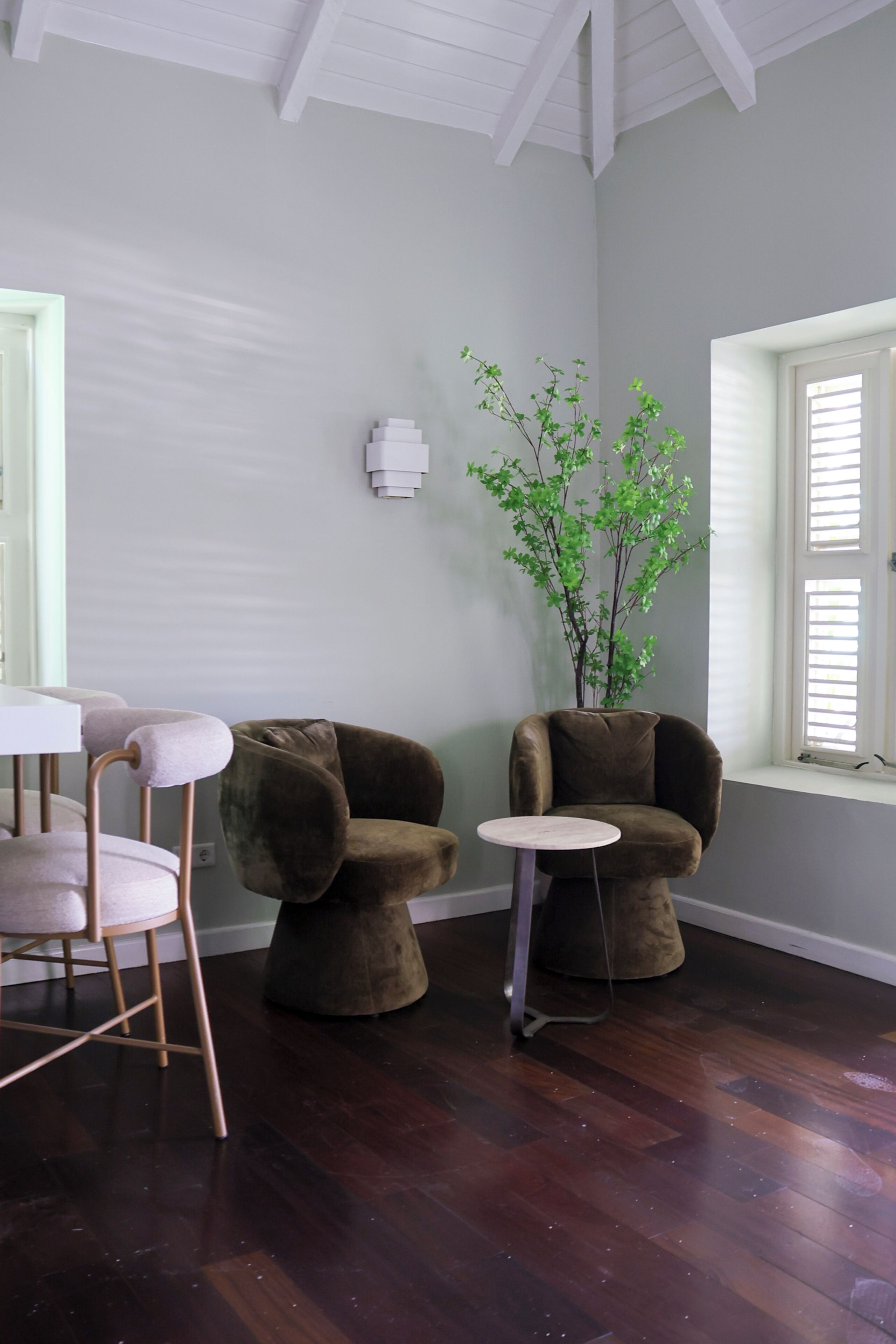 Una habitación con piso de madera oscura, pared blanca, una silla marrón, una mesita redonda pequeña y una ventana con persianas.