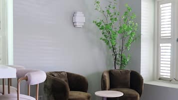 Una habitación con piso de madera oscura, pared blanca, una silla marrón, una mesita redonda pequeña y una ventana con persianas.