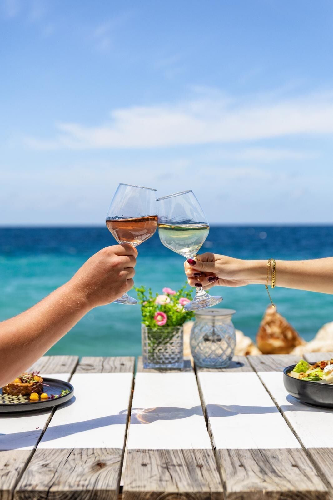 A wooden table with a glass of white wine and a glass of rosé wine being clinked together.