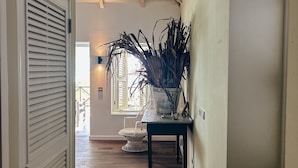 A hallway with wooden flooring, a white door with blinds, a potted plant, and a window with shutters. - BijBlauw Hotel (Willemstad)