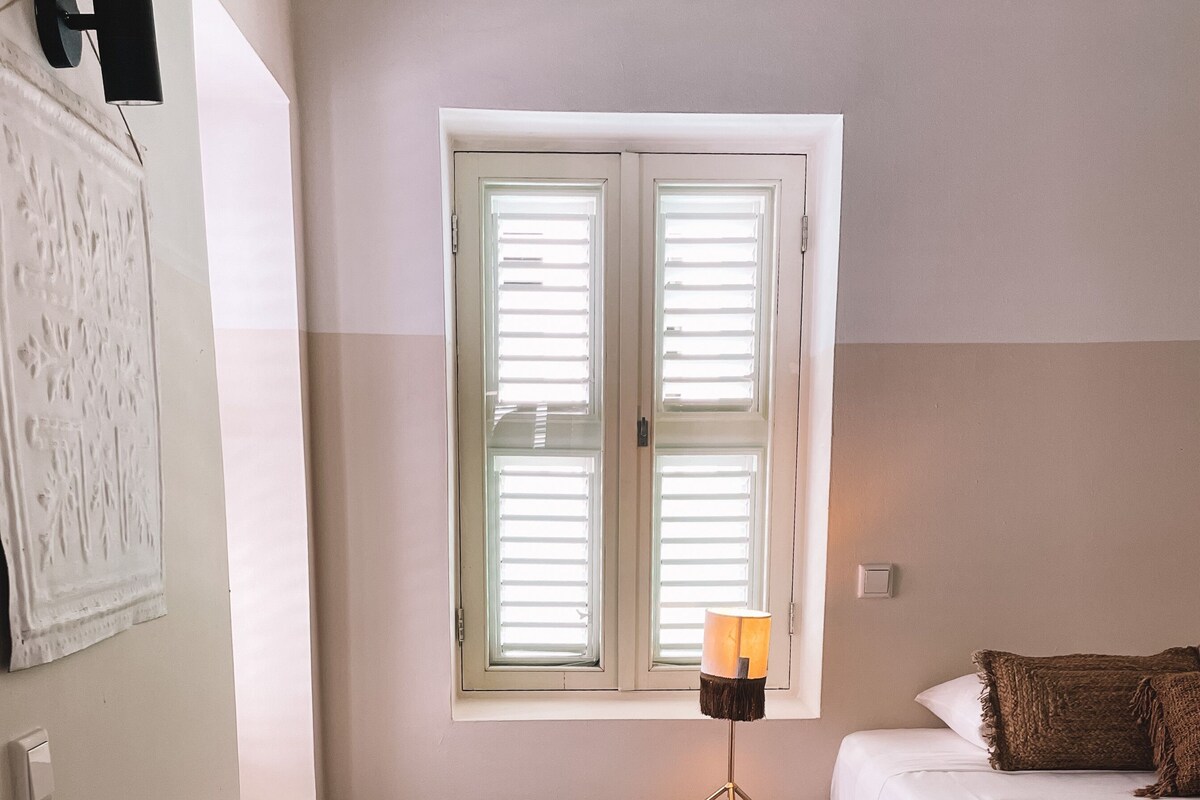 A bedroom with a bed, a small table, a lamp, and a window with blinds.