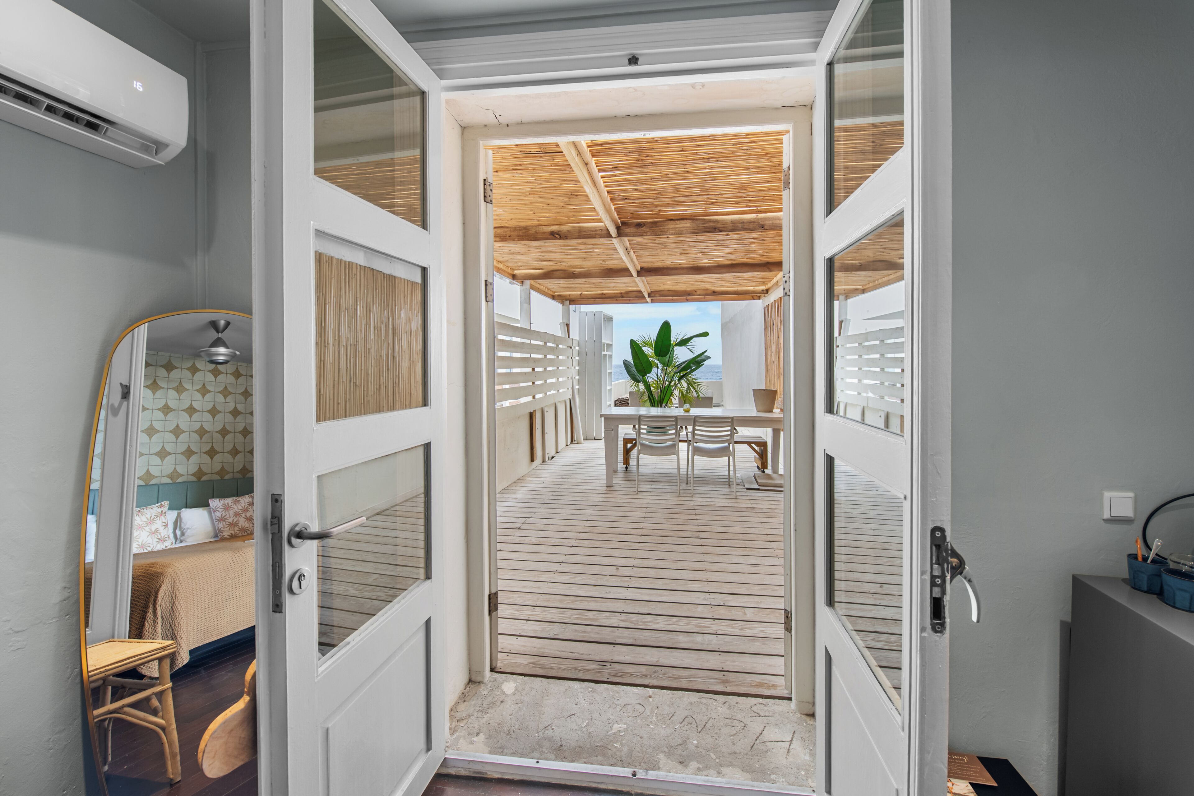 An open door leading to a balcony with a wooden floor, a table with chairs, and a potted plant.