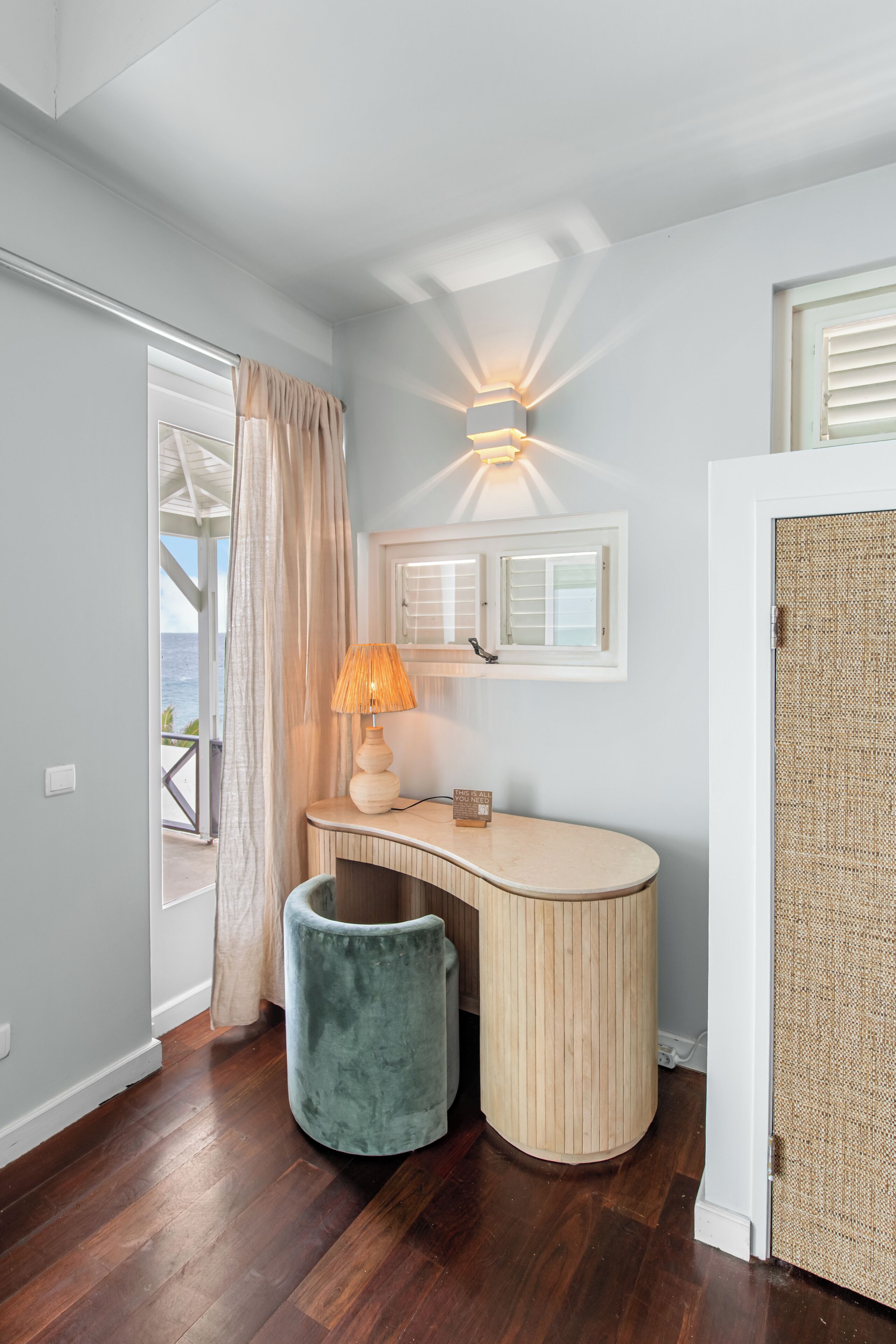 Une pièce comprenant un bureau en bois aux lignes courbes, une lampe, un tabouret rembourré, une fenêtre avec des volets et une porte à la texture tissée.