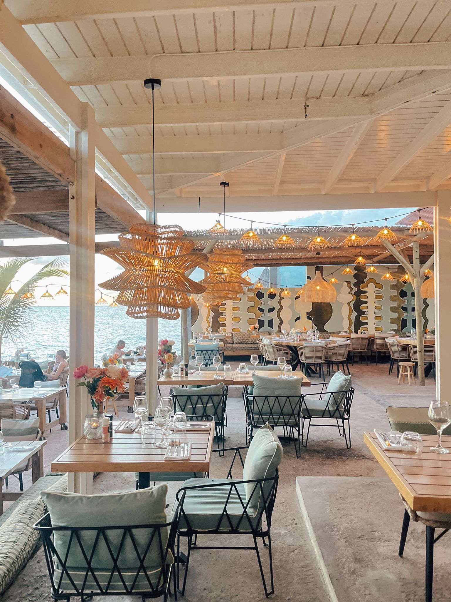 An outdoor restaurant with wooden tables and chairs, hanging wicker lamps, and a view of the sea.