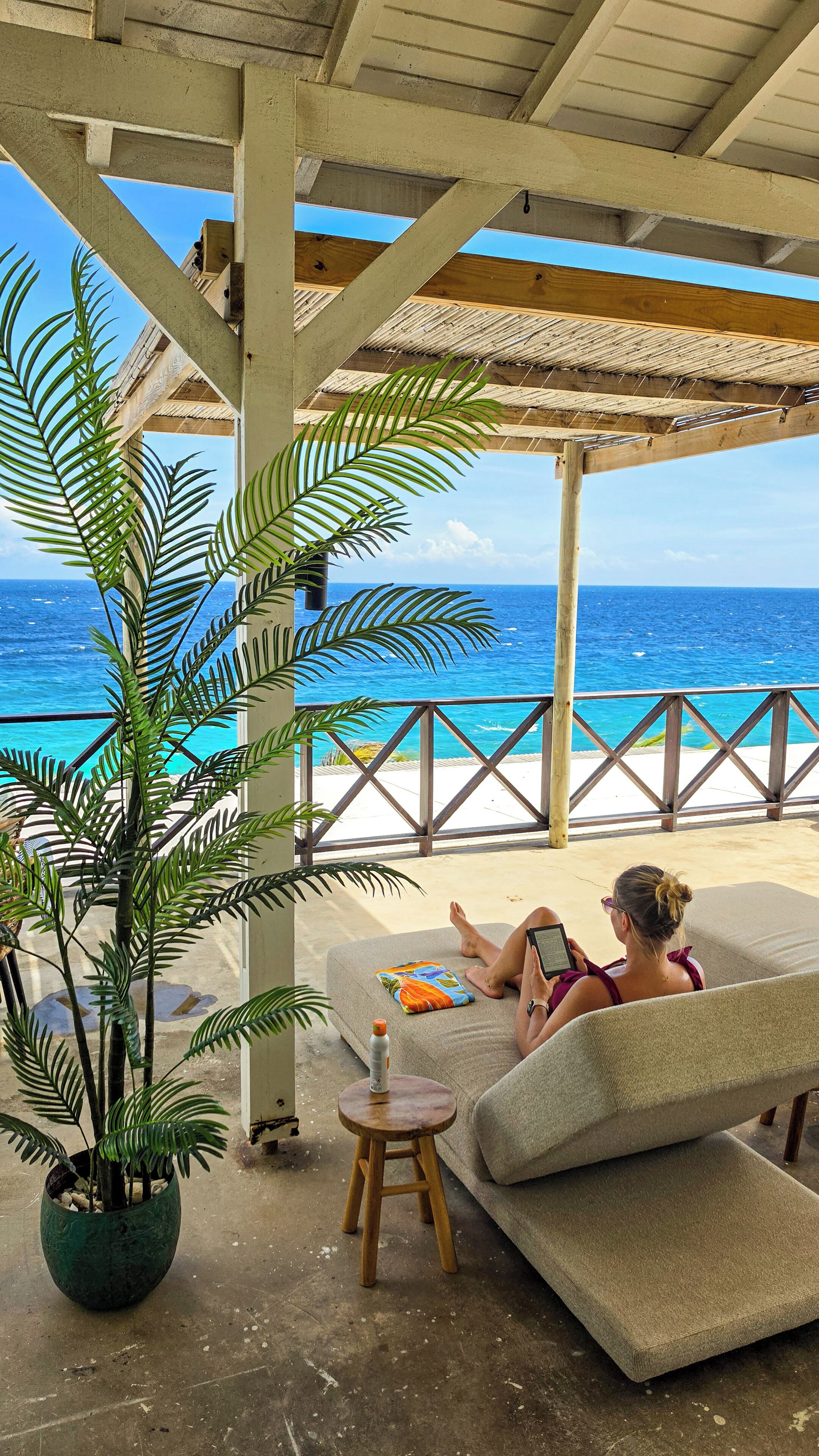 A covered outdoor seating area with a view of the ocean, a potted plant, and a small table with a bottle.