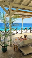 A covered outdoor seating area with a view of the ocean, a potted plant, and a small table with a bottle.