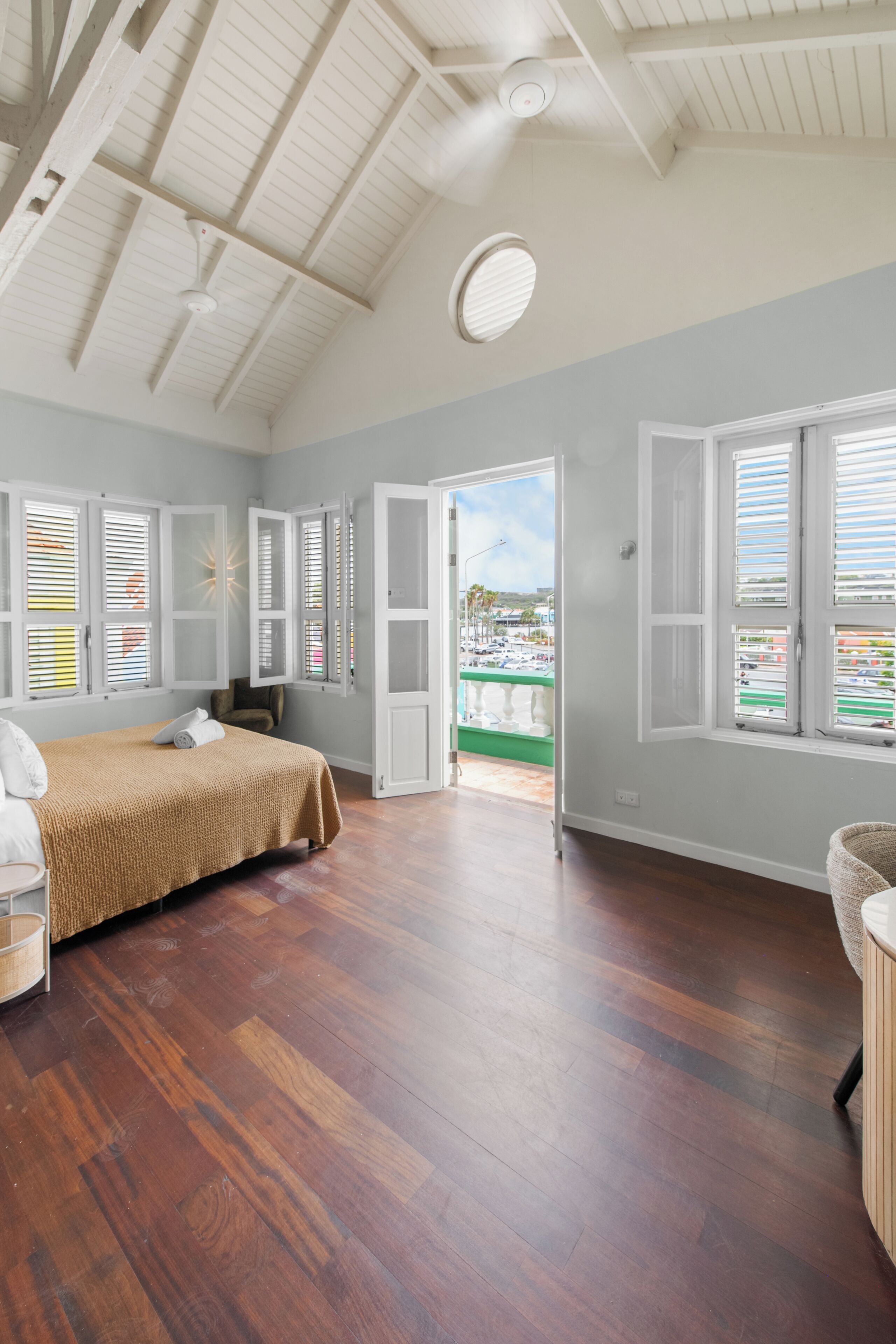 A spacious bedroom with a large bed, wooden flooring, and a view through an open door to the outside.