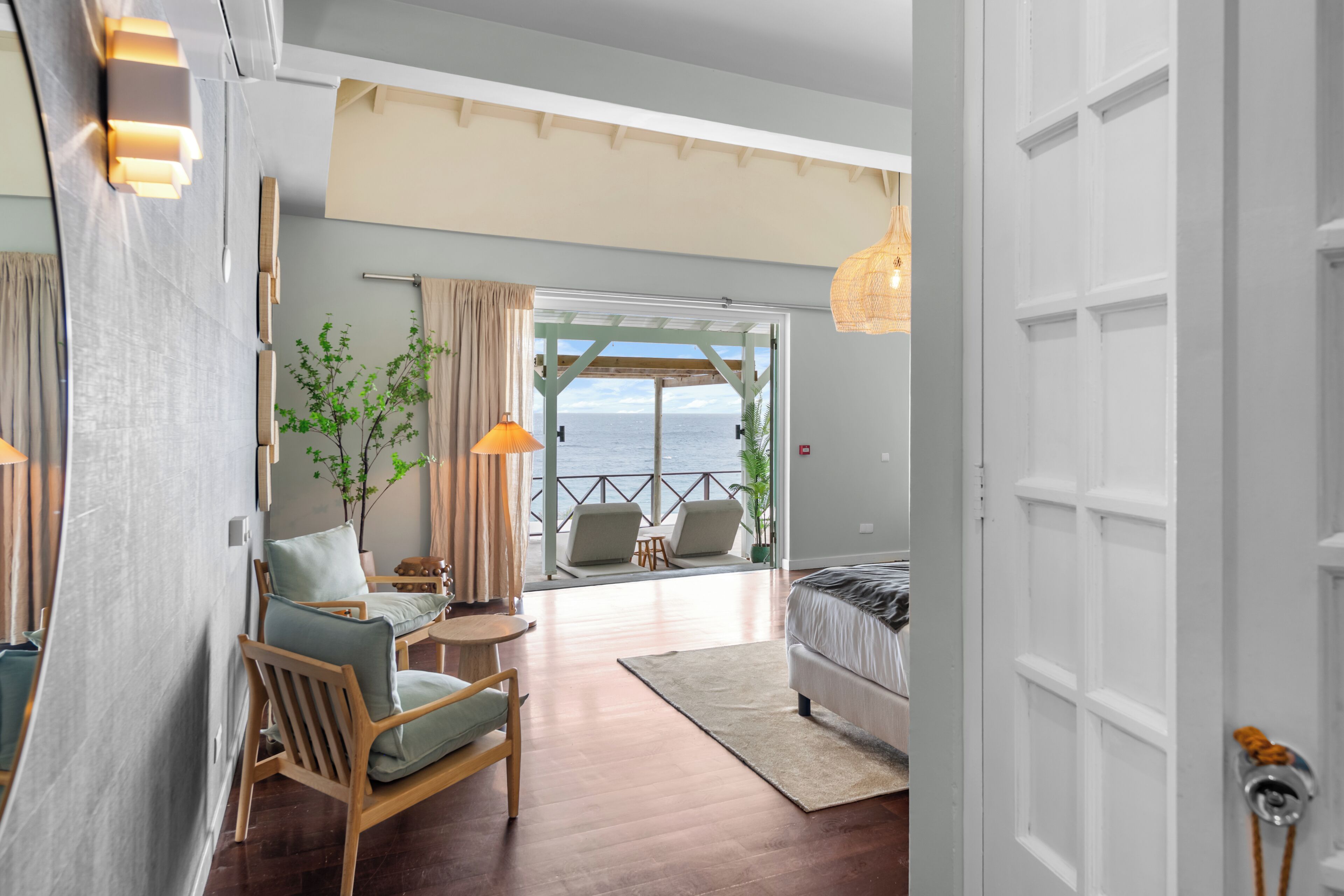 A room with a bed, a chair, a small table, and a view of the sea through an open door.