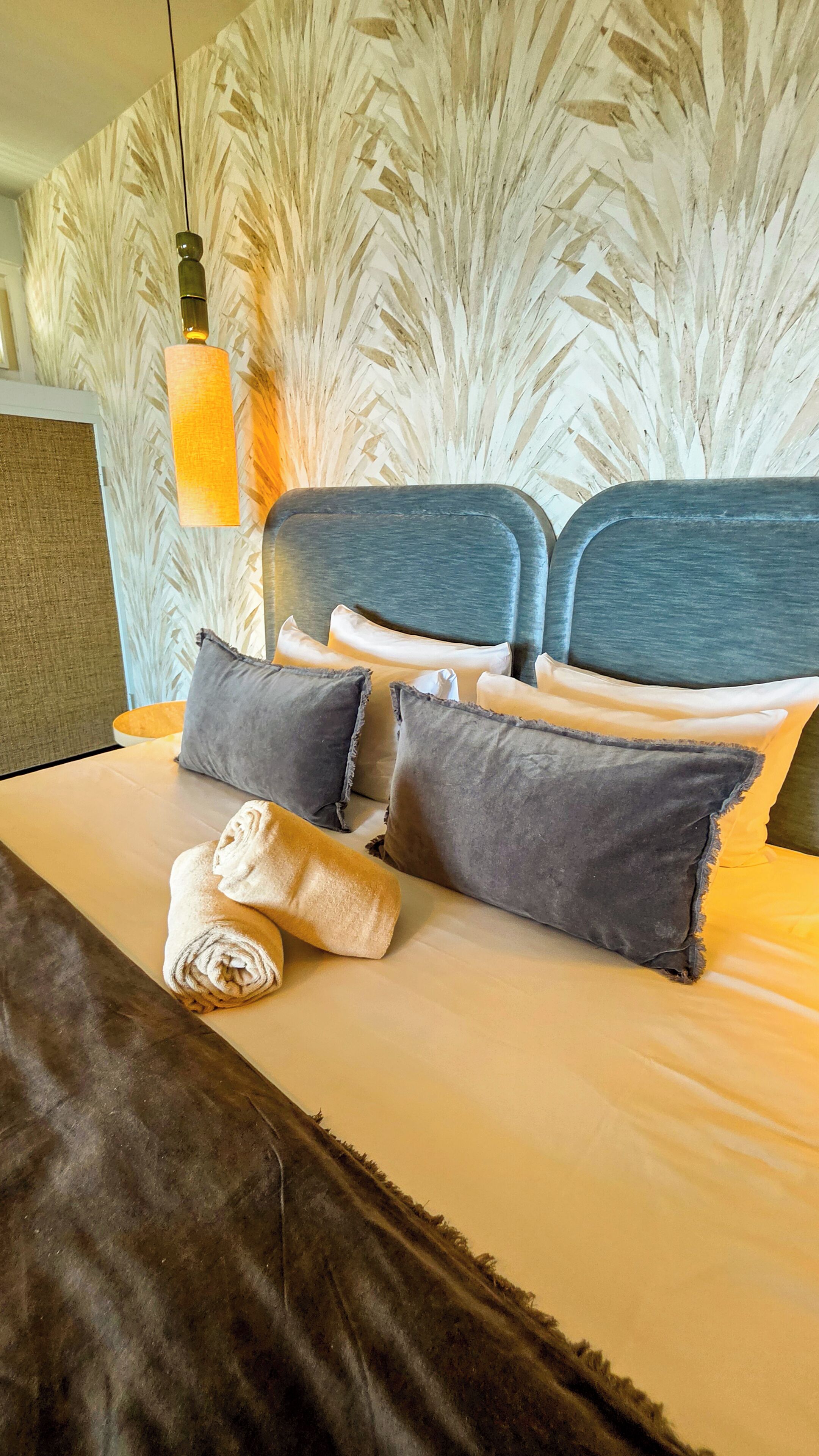 A neatly made bed with pillows and a rolled towel, a headboard with a textured pattern, and a hanging light fixture.