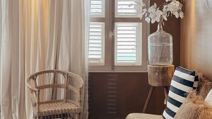 A room with a bed, a wicker chair, a hanging plant, and a window with shutters. - BijBlauw Hotel (Willemstad)