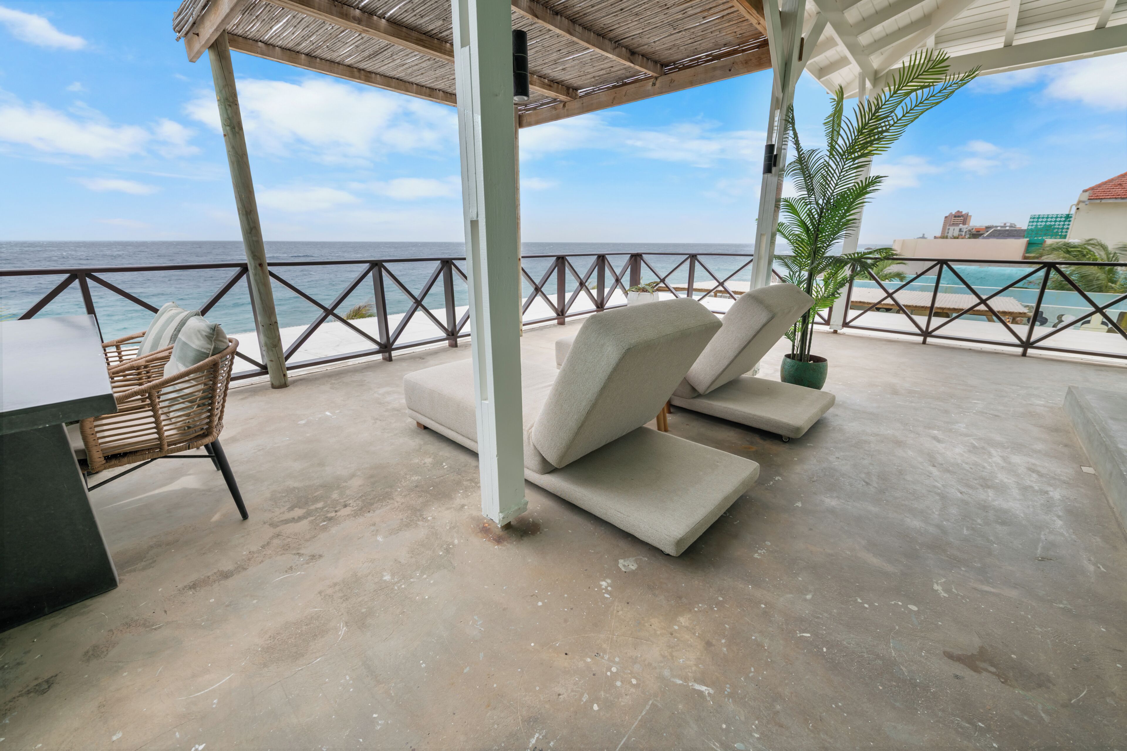 A covered outdoor seating area with a view of the ocean, a potted plant, and a pool in the background.